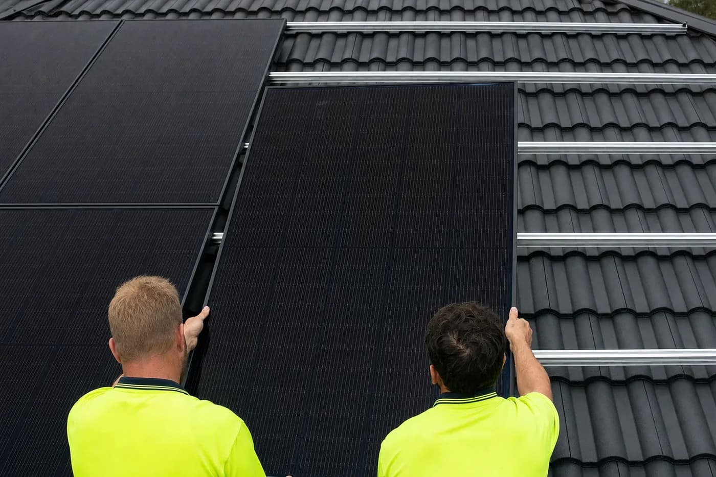 Two installers in hi-vis lowering an all-black solar panel onto the mounting rails of a half-finished rooftop array on a tiled roof