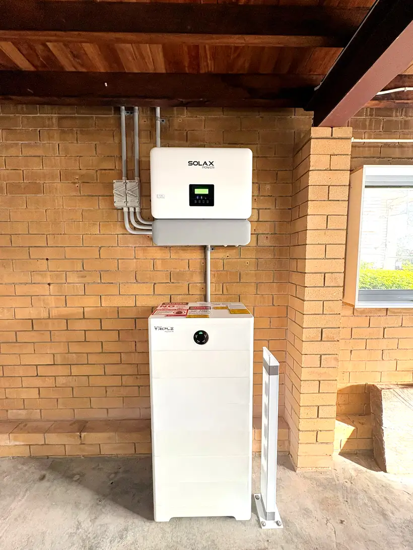 An inverter mounted on a brick wall above a floor-standing home battery, installed under a covered outdoor area
