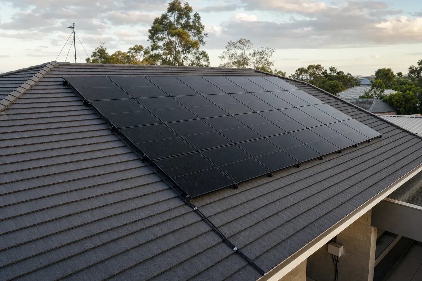 A finished array of black solar panels on a charcoal tiled roof, mounting rails and clamps visible along the lower edge and conduit running down to an isolator on the wall below