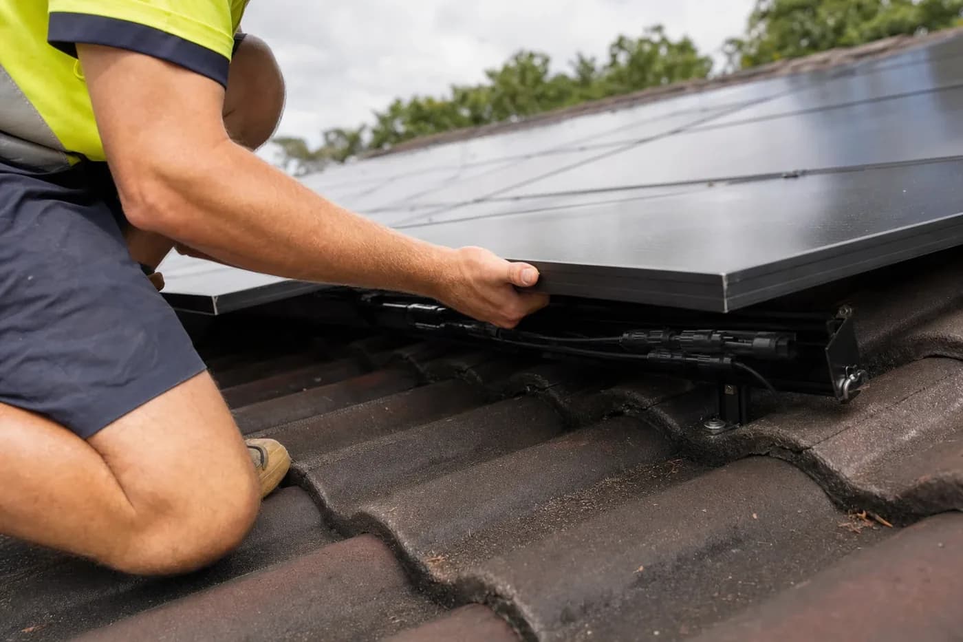 A technician in hi-vis kneeling on a tiled roof, lifting the lower edge of an all-black solar panel to check the DC connectors underneath it