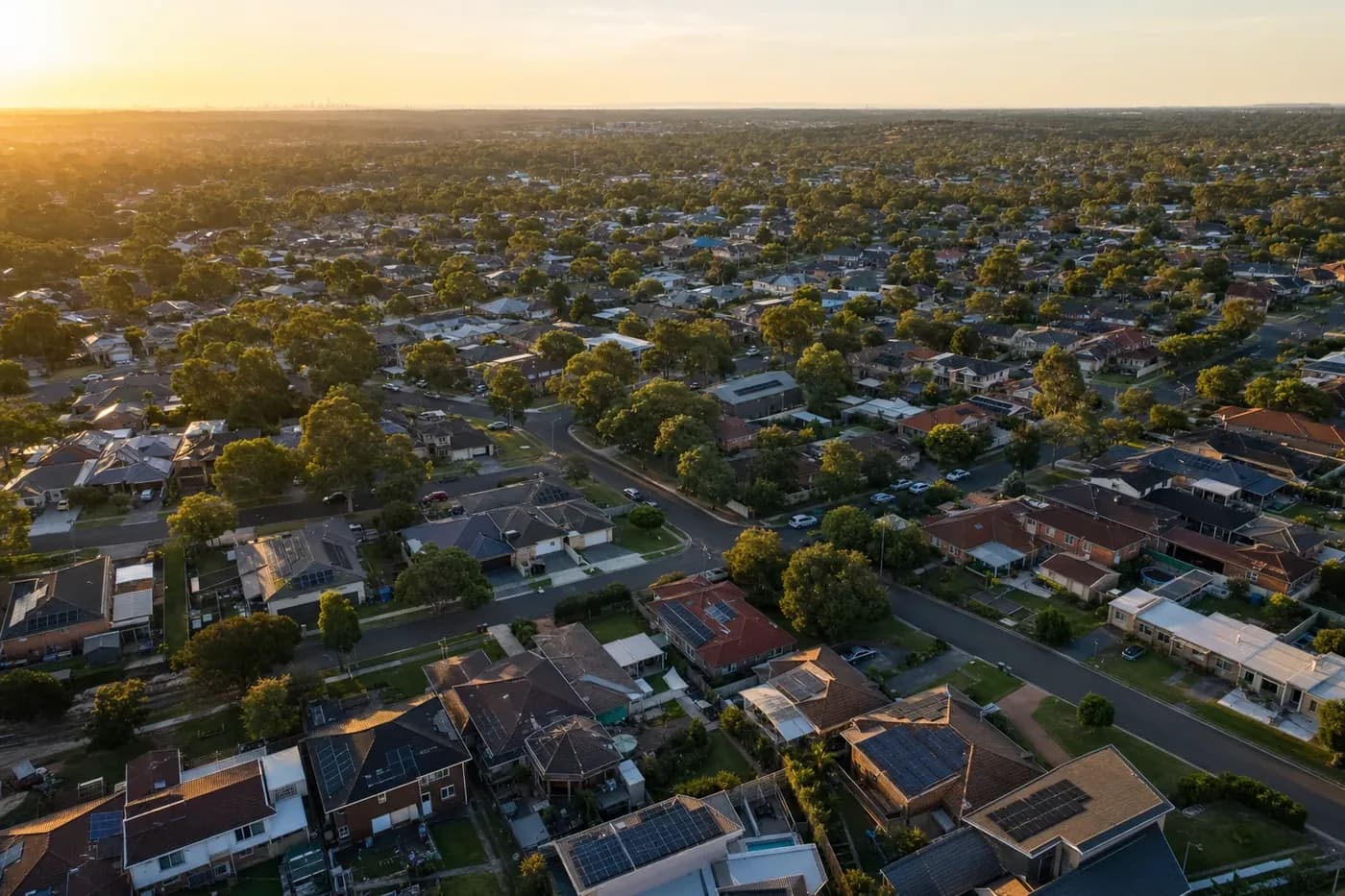 An aerial view across Western Sydney suburbia at golden hour, rows of tile and metal rooftops running to the horizon